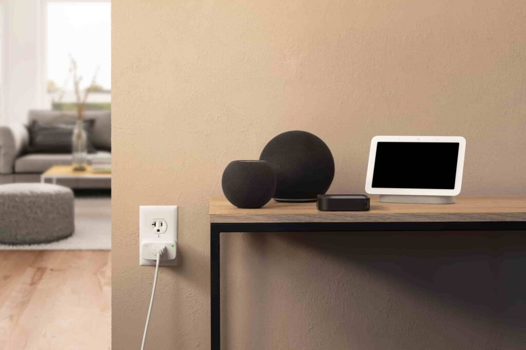 A white Eve Energy Matter smart plug inserted into a US wall outlet in a modern living room next to smart home speakers.